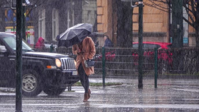A woman in a brown coat under an umbrella crosses a busy street in heavy rain