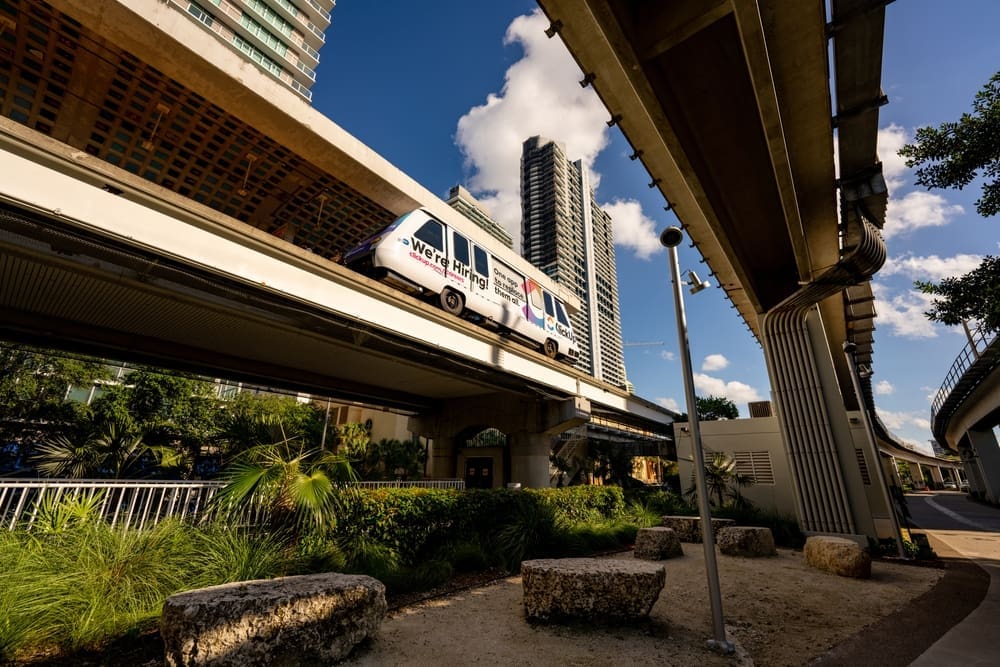The Miami Metromover train on its elevated track, passing over the landscaped Underline park area in Brickell, Miami, with skyscrapers and blue sky in the background.