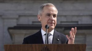 A close-up photograph of Mark Carney, a distinguished man with gray hair, wearing a dark suit and tie, speaking at a wooden podium outdoors.