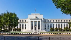 A daytime front view of the neoclassical Federal Reserve Board Building (Eccles Building) in Washington, D.C., featuring grand columns, a grand entrance, and an eagle statue on top, set against a clear blue sky.
