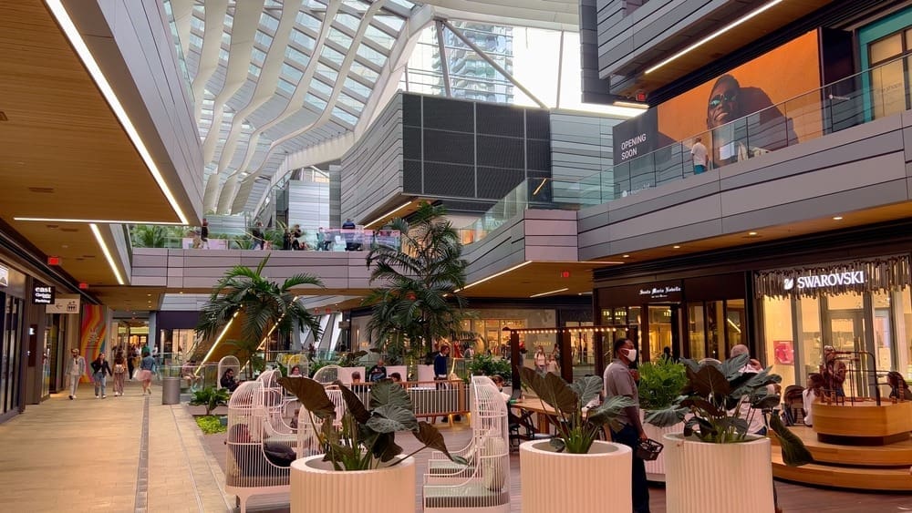 The bright, modern interior of Brickell City Centre, a multi-story shopping mall with walkways, a curved architectural ceiling, luxury stores like Swarovski, and lush tropical plants.
