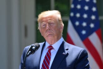 President Donald Trump stands at an outdoor podium with two microphones, with a large American flag in the background.