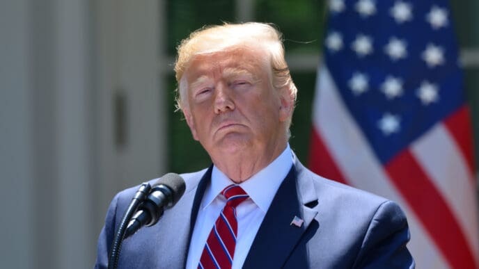 President Donald Trump stands at an outdoor podium with two microphones, with a large American flag in the background.