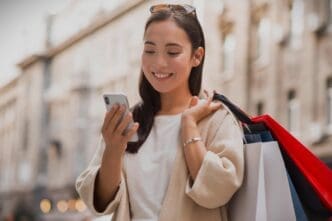 Smiley Chinese woman looking at smartphone outdoors while holding shopping bags