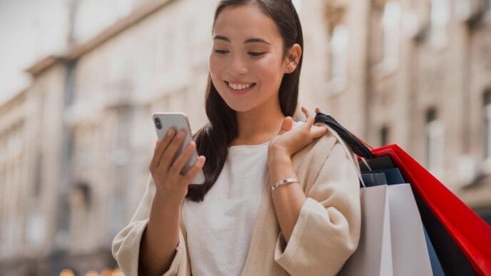Smiley Chinese woman looking at smartphone outdoors while holding shopping bags