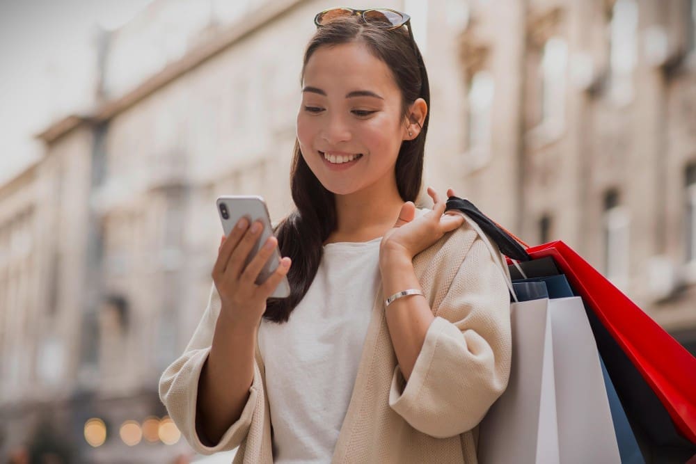 Smiley Chinese woman looking at smartphone outdoors while holding shopping bags