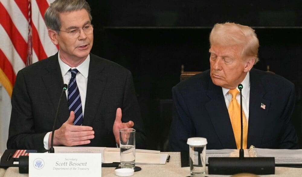 Scott Bessent, with a name card identifying him as "Secretary Scott Bessent, Department of the Treasury," speaks at a table while Donald Trump listens intently. They are in a formal meeting setting, with a U.S. flag visible in the background.