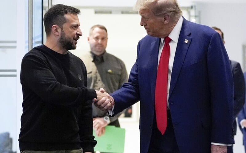 Volodymyr Zelenskyy and President Donald Trump shake hands in an indoor setting, with a serious and focused expression.