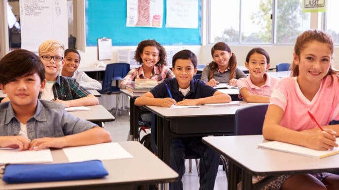 Smiling elementary school kids sitting at desks in classroom. By Shutterstock.com - Monkey Business Images
