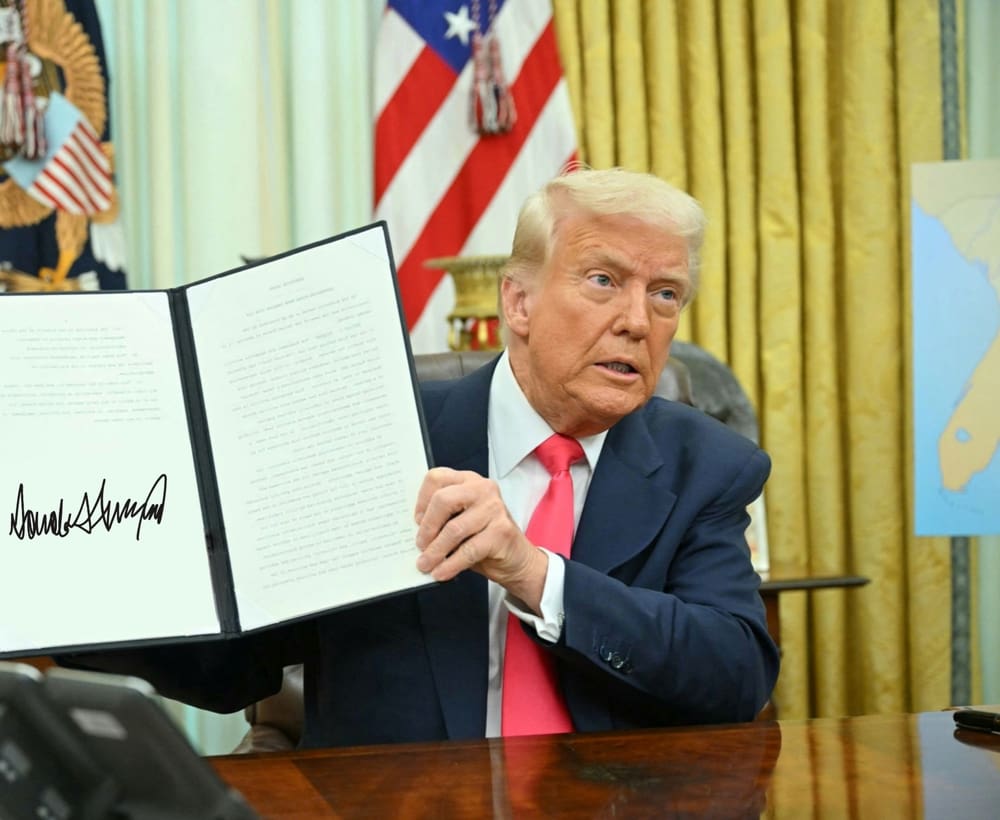 President Donald Trump sits at the Resolute Desk in the Oval Office, holding up a signed executive order for the camera.
