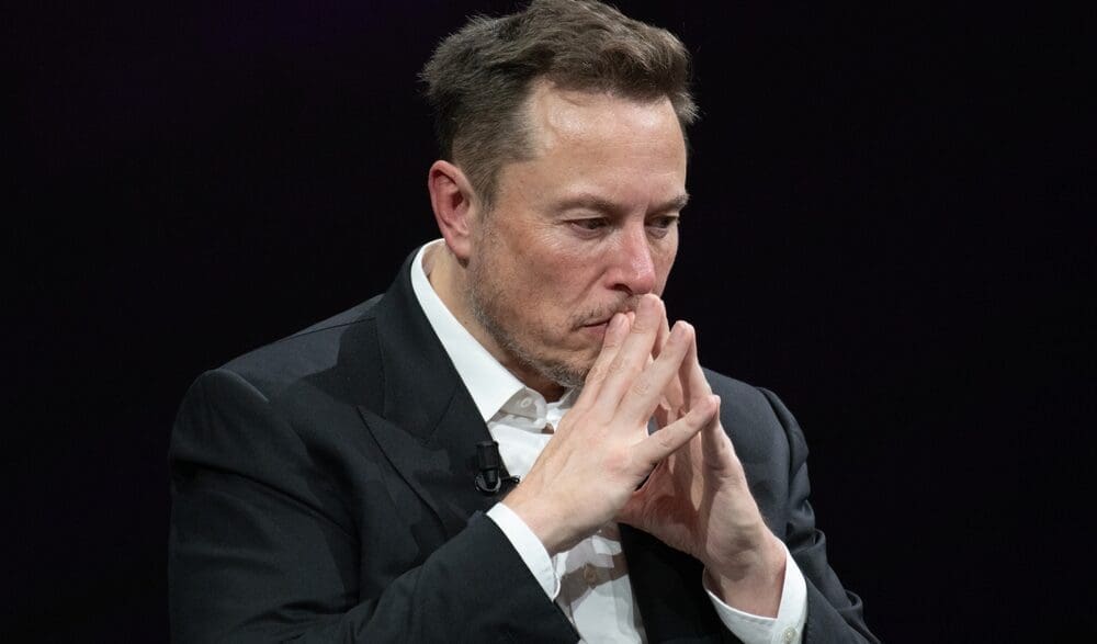 A close-up portrait of Elon Musk looking downwards with a pensive expression, his hands clasped together near his mouth, against a dark, plain background.