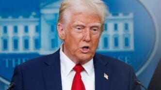 A close-up portrait of Donald Trump speaking at a podium, wearing a dark suit and red tie, with a blurred blue backdrop featuring a stylized White House building.