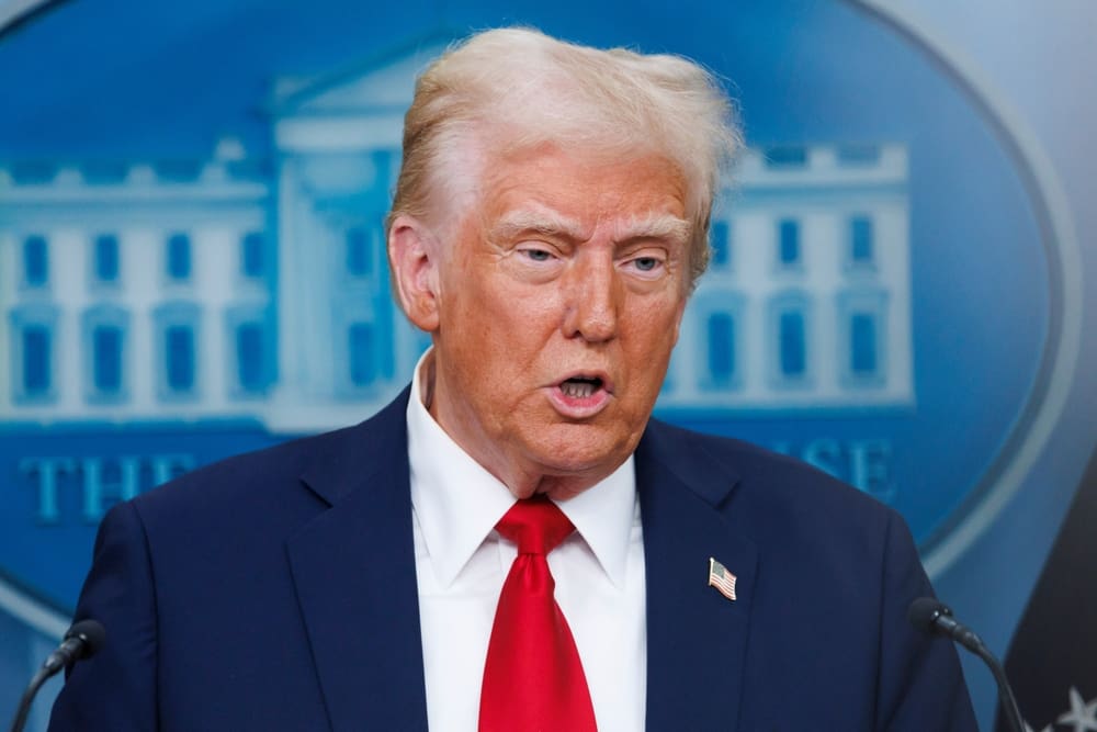 A close-up portrait of Donald Trump speaking at a podium, wearing a dark suit and red tie, with a blurred blue backdrop featuring a stylized White House building.