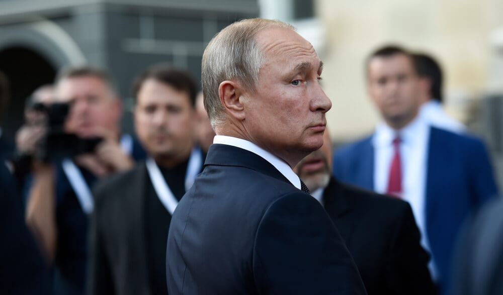 A profile shot of Russian President Vladimir Putin, wearing a dark suit, looking intently to his right, with a blurred crowd and a photographer in the background.