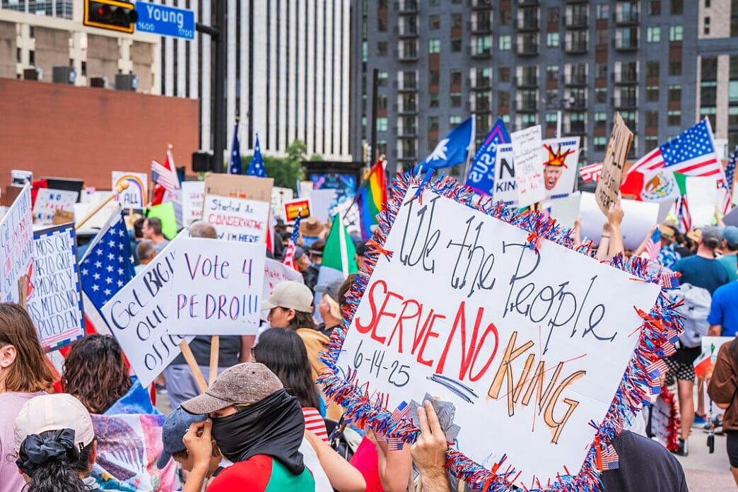 A dense crowd of protesters on a city street in Dallas, Texas, holding various signs and flags. A prominent handmade sign in the foreground reads, "We the People SERVE NO KING" and is dated "6-14-25".