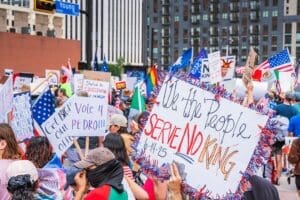 A dense crowd of protesters on a city street in Dallas, Texas, holding various signs and flags. A prominent handmade sign in the foreground reads, "We the People SERVE NO KING" and is dated "6-14-25".