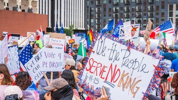 A dense crowd of protesters on a city street in Dallas, Texas, holding various signs and flags. A prominent handmade sign in the foreground reads, "We the People SERVE NO KING" and is dated "6-14-25".