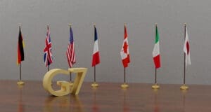 A 3D rendering of a golden "G7" logo on a wooden table, with the seven national flags of the G7 member countries standing in a row behind it.