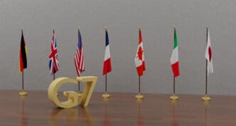A 3D rendering of a golden "G7" logo on a wooden table, with the seven national flags of the G7 member countries standing in a row behind it.