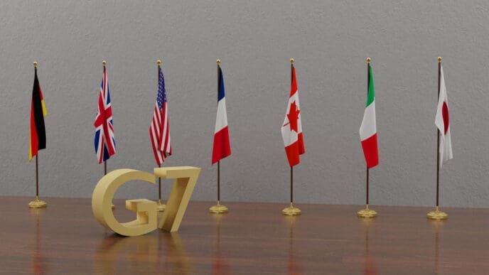 A 3D rendering of a golden "G7" logo on a wooden table, with the seven national flags of the G7 member countries standing in a row behind it.