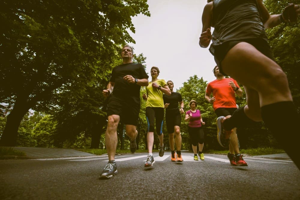 A low-angle action photograph of a diverse group of people in athletic gear running towards the camera on a paved path that is lined with large, green trees.