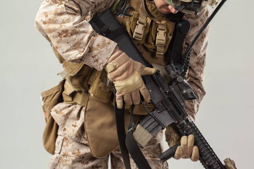 A close-up, mid-section view of a U.S. Marine in desert camouflage and full combat gear, holding a rifle against a neutral studio background.