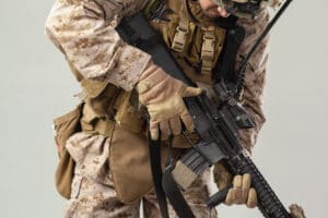 A close-up, mid-section view of a U.S. Marine in desert camouflage and full combat gear, holding a rifle against a neutral studio background.