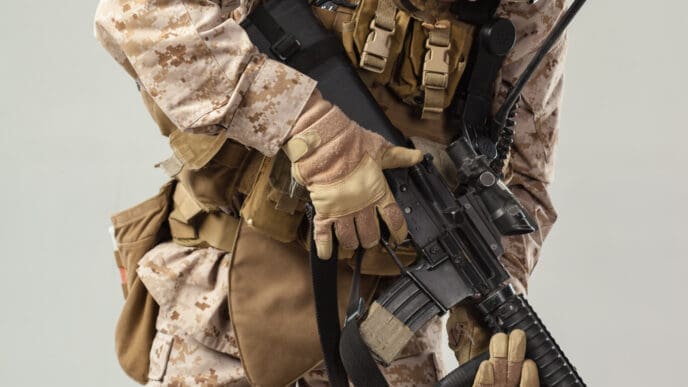 A close-up, mid-section view of a U.S. Marine in desert camouflage and full combat gear, holding a rifle against a neutral studio background.
