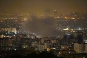 A nighttime photograph of the Tehran cityscape showing a large plume of smoke rising from among the illuminated buildings, reportedly following an Israeli attack on June 13, 2025.