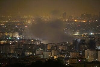 A nighttime photograph of the Tehran cityscape showing a large plume of smoke rising from among the illuminated buildings, reportedly following an Israeli attack on June 13, 2025.