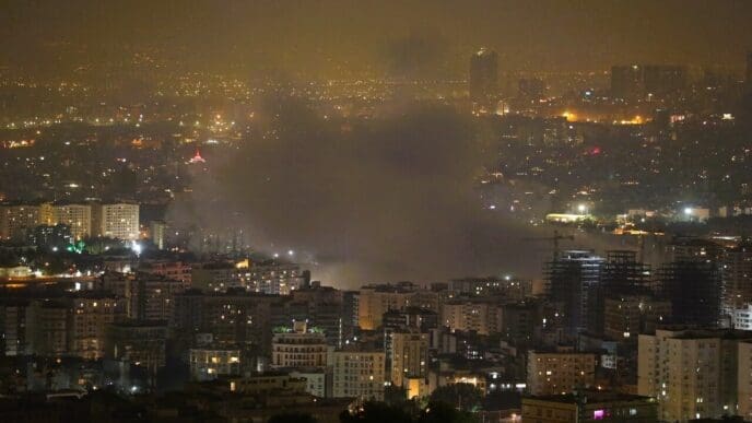 A nighttime photograph of the Tehran cityscape showing a large plume of smoke rising from among the illuminated buildings, reportedly following an Israeli attack on June 13, 2025.