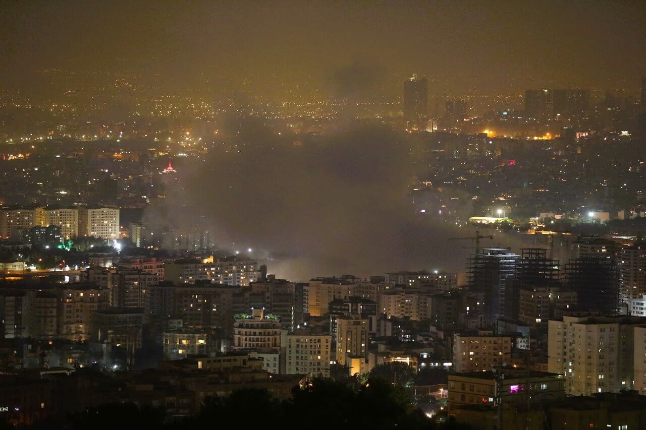 A nighttime photograph of the Tehran cityscape showing a large plume of smoke rising from among the illuminated buildings, reportedly following an Israeli attack on June 13, 2025.