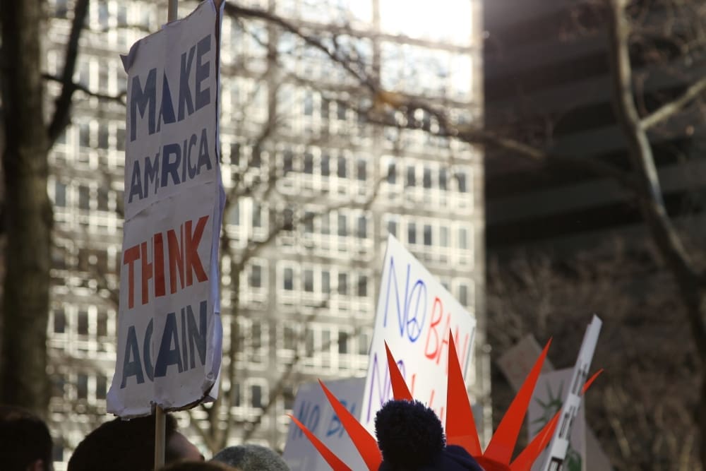 A low-angle view of protest signs at a rally, with one prominent banner reading "Make America Think Again" in blue and red letters, held up in front of a large city building.