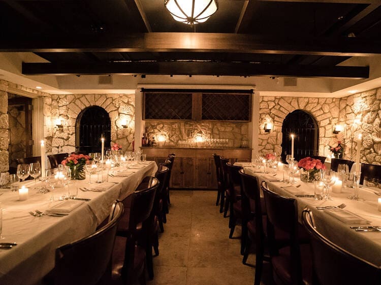 The dimly lit interior of Ariete restaurant in Coconut Grove, showing two long dining tables set with white tablecloths, candles, and wine glasses, all framed by rustic stone walls.