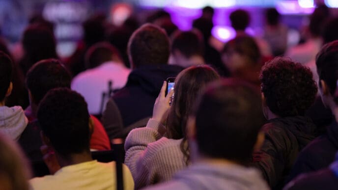 A view from behind a seated audience in a dimly lit hall. One person in the foreground holds up a smartphone, seemingly recording the stage, which is illuminated with bright purple lights.