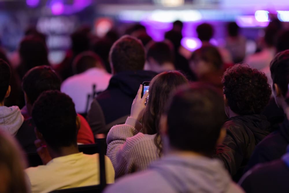 A view from behind a seated audience in a dimly lit hall. One person in the foreground holds up a smartphone, seemingly recording the stage, which is illuminated with bright purple lights.