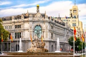 The grand, ornate building of the Bank of Spain in Madrid, with the elaborate Cibeles Fountain featuring lion sculptures in the foreground. Spanish flags fly around the fountain under a partly cloudy sky.