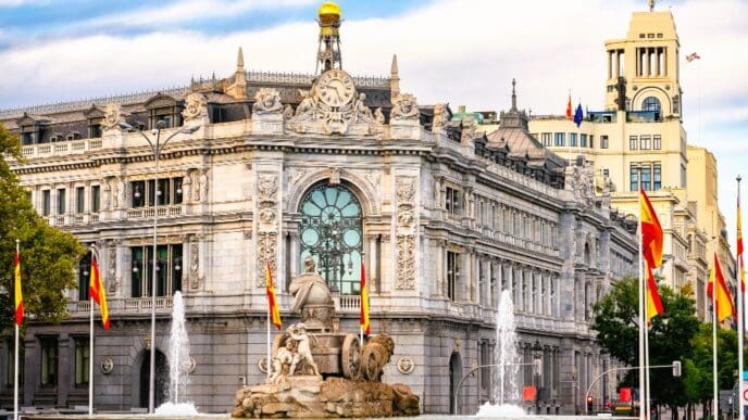 The grand, ornate building of the Bank of Spain in Madrid, with the elaborate Cibeles Fountain featuring lion sculptures in the foreground. Spanish flags fly around the fountain under a partly cloudy sky.