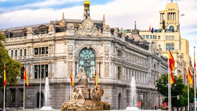 The grand, ornate building of the Bank of Spain in Madrid, with the elaborate Cibeles Fountain featuring lion sculptures in the foreground. Spanish flags fly around the fountain under a partly cloudy sky.