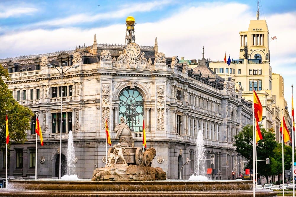 The grand, ornate building of the Bank of Spain in Madrid, with the elaborate Cibeles Fountain featuring lion sculptures in the foreground. Spanish flags fly around the fountain under a partly cloudy sky.