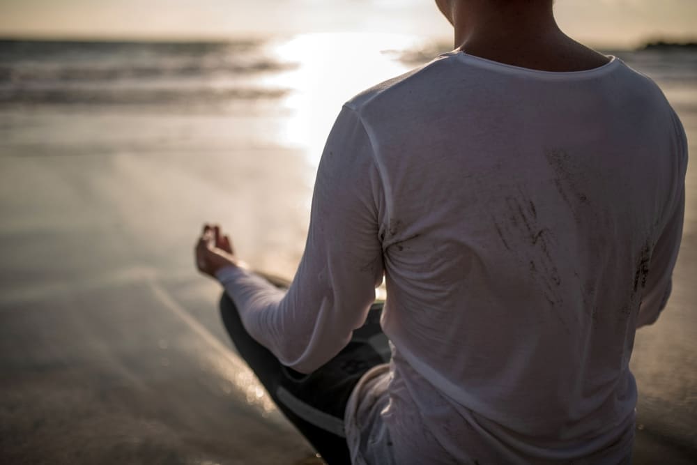A person is seen from behind, sitting in a cross-legged meditation pose on a wet beach at sunset or sunrise, with their hands in a mudra as the sun's reflection shimmers on the water.