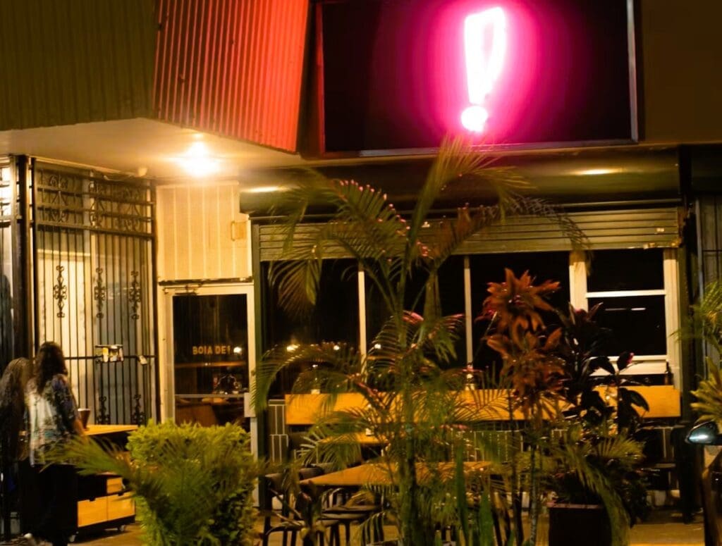 The nighttime exterior of Boia De Italian restaurant in Miami, featuring its iconic glowing pink neon exclamation mark sign above the entrance, with outdoor patio seating and plants.