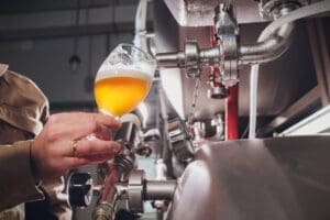 A close-up of a person's hand holding a stemmed glass, pouring a hazy, golden beer directly from a tap on a large stainless steel brewing tank inside a brewery.