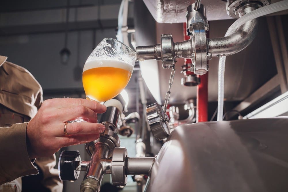 A close-up of a person's hand holding a stemmed glass, pouring a hazy, golden beer directly from a tap on a large stainless steel brewing tank inside a brewery.