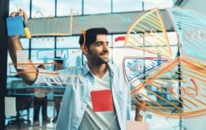 A smiling businessman, who appears to be of South Asian or Middle Eastern descent, stands and writes a business plan with colorful charts and graphs on a transparent board.