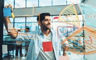 A smiling businessman, who appears to be of South Asian or Middle Eastern descent, stands and writes a business plan with colorful charts and graphs on a transparent board.