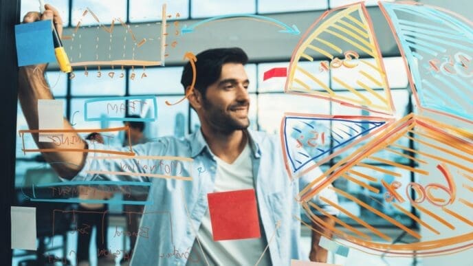 A smiling businessman, who appears to be of South Asian or Middle Eastern descent, stands and writes a business plan with colorful charts and graphs on a transparent board.