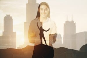 A double exposure image featuring a smiling professional woman with her arms crossed, blended with a city skyline at sunset. A second silhouette of a person in a victory pose on a mountaintop is visible within her figure.