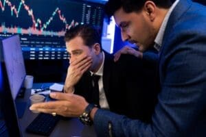Two male financial professionals in suits look stressed in front of multiple computer screens displaying volatile stock charts. One man covers his mouth in dismay, while the other places a comforting hand on his shoulder.