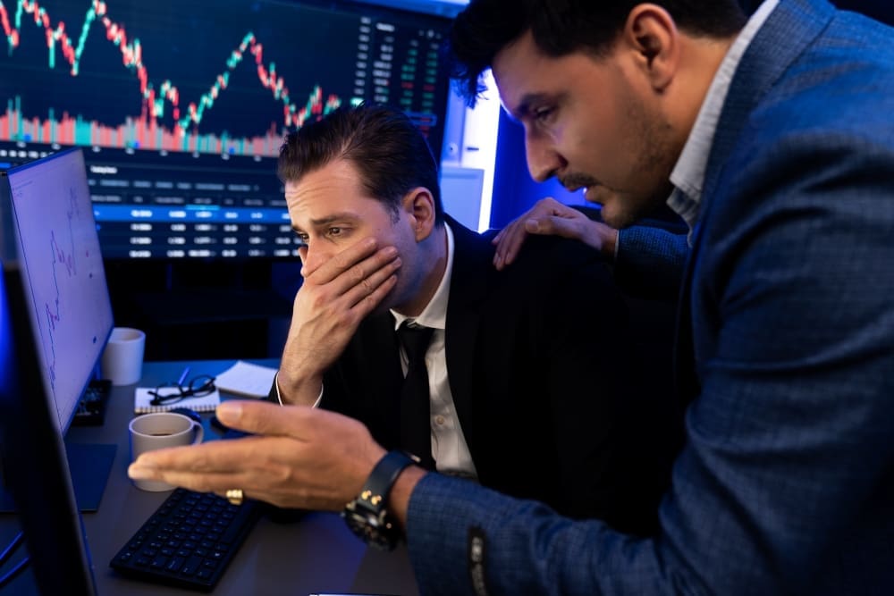 Two male financial professionals in suits look stressed in front of multiple computer screens displaying volatile stock charts. One man covers his mouth in dismay, while the other places a comforting hand on his shoulder.